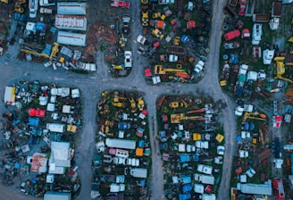 Aerial view of an extensive junkyard with various old and abandoned vehicles, including cars, trucks, and trailers. The area is divided into multiple sections by dirt paths, and various metal parts and machinery are scattered throughout. The vehicles and parts are in different stages of disrepair, with rust and wear evident.